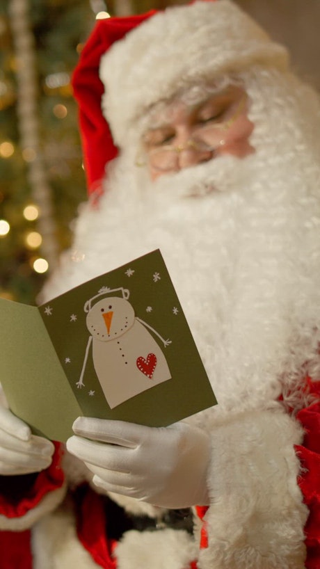 A charming Santa Claus wearing reading glasses peruses trough a hand crafted letter adorner with a snow man in the cover.