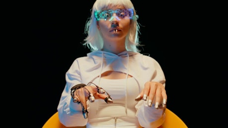 A futuristic woman in white attire and a hand sci fi device on her hands delicately moves her metal fingers.