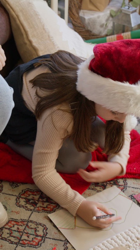 A girl wearing a red Santa Claus hat working on a Christmas greeting card on the floor.