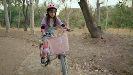 A liitle girl with pink helmet rides a bike in the park.