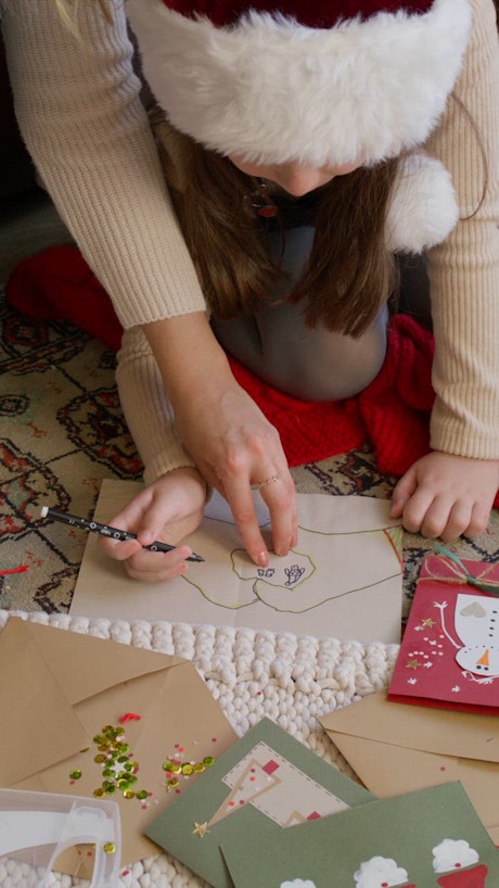 A little girl positioned comfortably on the floor receives help from an adult with the Christmas greeting card decoration.