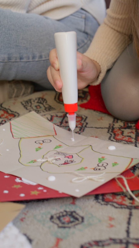 A little kid adds little drops of white glue as decoration over a hand-crafted letter to Santa Claus.
