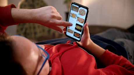 A man lying on the bed scrolling food pictures on his phone.
