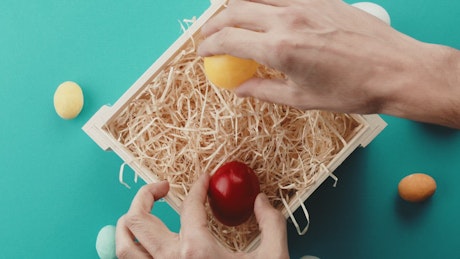 A man's hand placing colored Easter Eggs in a basket.