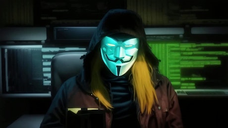A mysterious person with a mask of V from Vendetta is sitting with computer code screens in the background.