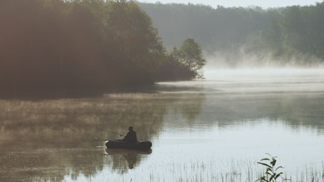 A person fishing on a boat on a misty early morning.
