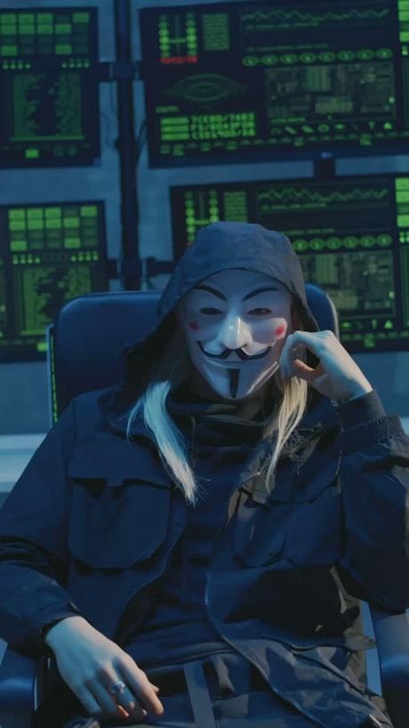 A person wearing an anonymous mask sits relaxed on a chair in front of a bank of computer screens.
