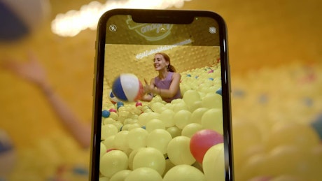 A smartphone records a young happy woman playing in the plastic ball pit pool.