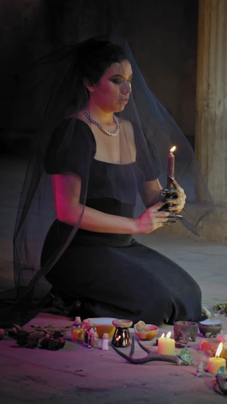 A vertical shot creepy looking a woman with blank eyes and a dark veil is whispering mystical words in front of an arrangement of ritual objects.