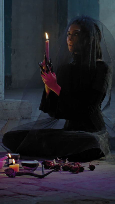 A witch sitting on the ground prays while holding a candle.