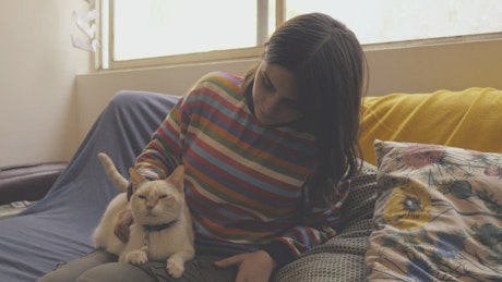 A woman sits on a couch and pets a cat.