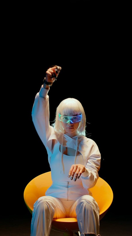 A woman with white hair and a sleek white attire sits on a yellow chair wearing sci fi devices on her eyes and her hands to control the digital realm.