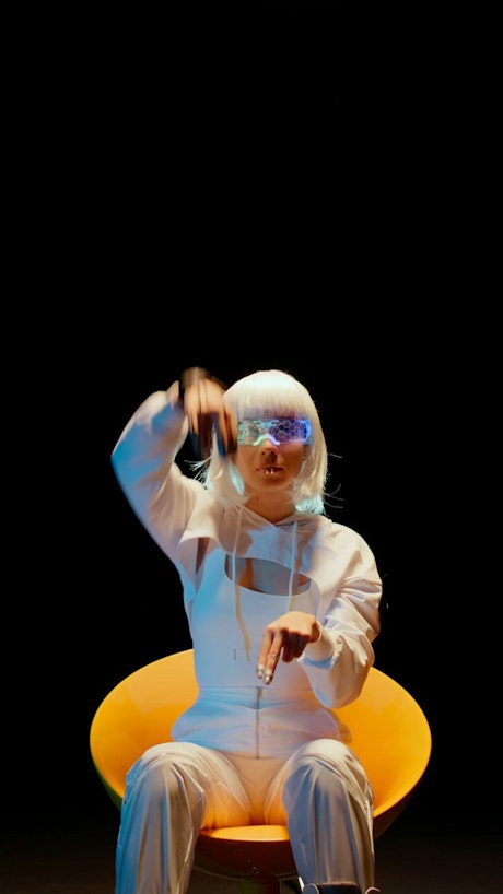 A woman with white hair and white suit, sits on a vibrant yellow chair moving her hands up and down.
