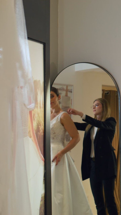 A young beautiful bride stands in front of the mirror as she tries on brand new white wedding gown.
