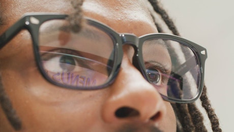 A young black man with dreadlocks and glasses looking at the computer screen.