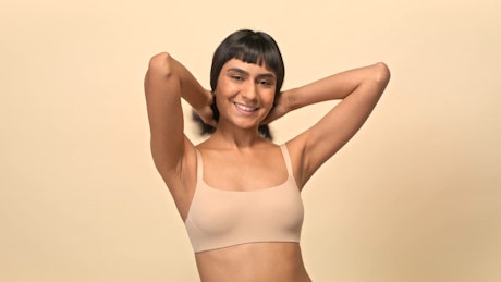 A young brunette woman clad in beige underwear strikes poses to the camera over a beige photo studio background.