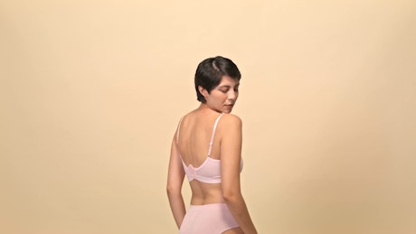 A young brunette woman in pink underwear with her back facing to the camera slowly turns over a beige photo studio backdrop.