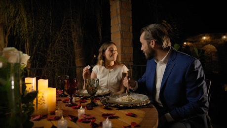 A young couple celebrates Valentine's Day sharing pasta and wine in a table adorned with rose petals and candles.