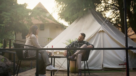 A young couple sitting outside a white fabric camping tent make a toast with wine glasses.