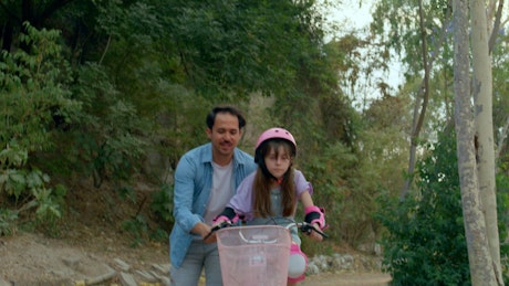 A young girl focuses on riding a bike while her father helps her.