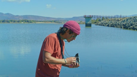 A young man adjust an old photography camera by the lake.