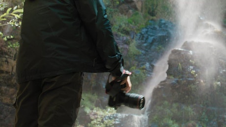 A young man raises his professional camera to take a picture of a waterfall.