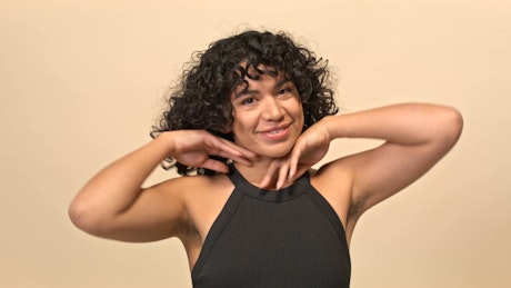 A young natural brunette woman plays with her curly black hair over a beige photo studio background.