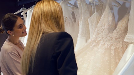 A young sales woman shows bride dresses to a bride to be young woman at the store wardrobe.