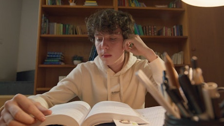 一个年轻的学生在家里读一本书，书的背景是一个装满书的书架