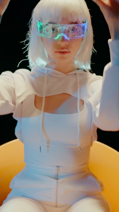 A young woman in white attire and a futuristic sci fi VR glasses concentrates on an unseen digital task moving her hands.