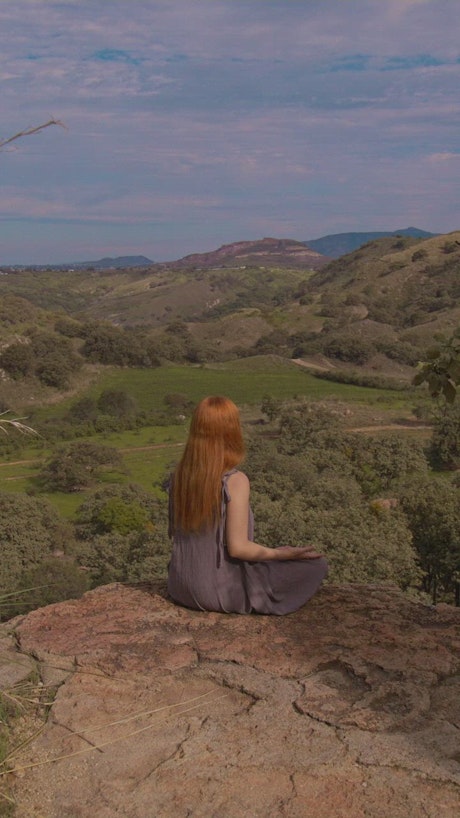 A young woman meditating high up in nature sitting over a huge rock with mesmerizing mountains in the background.