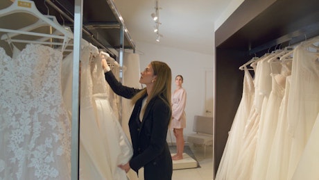 A young woman select an exquisite white bride dress with a young bride to be waiting for the dress in the back of the changing room.
