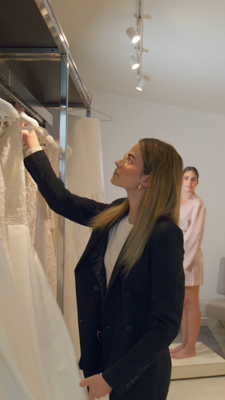 A young woman takes out a beautiful with wedding gown from the wardrobe to deliver it to the eager client waiting in front of the mirror.