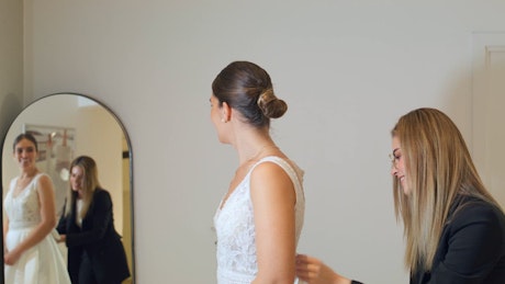 A young woman trying a beautiful white bride gown in front of a mirror with a young woman asisting her on her back.