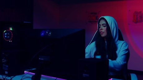 A young woman wearing a hoodie puts on her headphones to start playing video games in front of the pc rig.