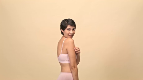 A young woman with black short hair and pink underwear stretches her hands and poses to the camera against a photo studio backdrop.