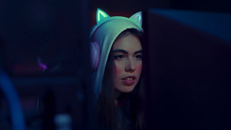 A young woman with white headphones adorned with cute feline ears smiles a rejoice in front of the pc rig.