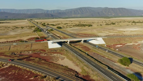 Aerial view of a highway overpass with a big red transport truck traversing the asphalt on a sunny day, arid landscape in the background.