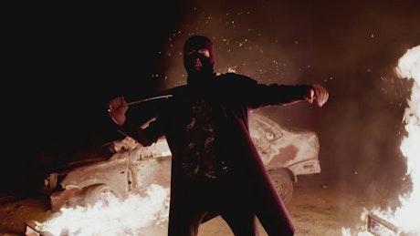 Angry vandal stands in front of a burning car during civil unrest.