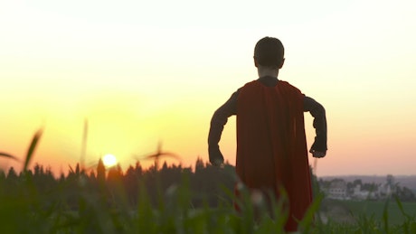 Boy dressed with a superhero red cape standing in a field.
