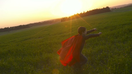 Boy runs through the countryside wearing a superhero cape.