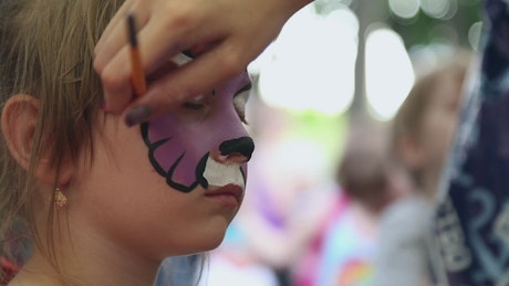 Child gets their face painted as a purple cat.