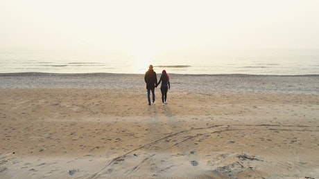 Couple holding hands walking on beach towards sea.