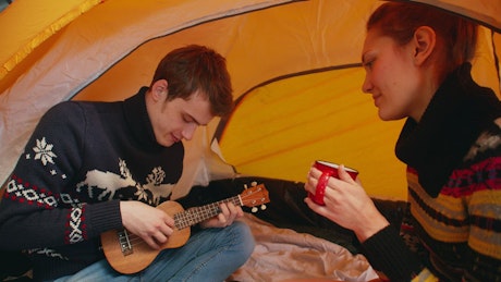 Couple sitting in a tent while camping playing the ukulele.