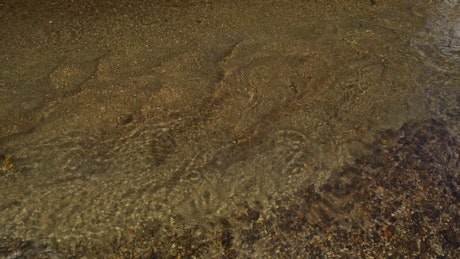 Crystal clear water gently passing by the riverbed full of sand and small rocks.