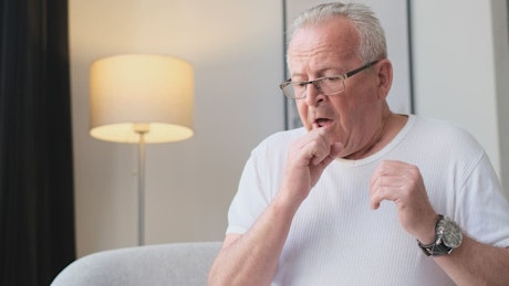 Elderly adult sitting on couch with headache and coughing.