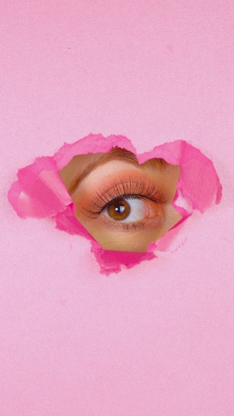 Eye of a woman peeking out of the hole of a pink paper screen.