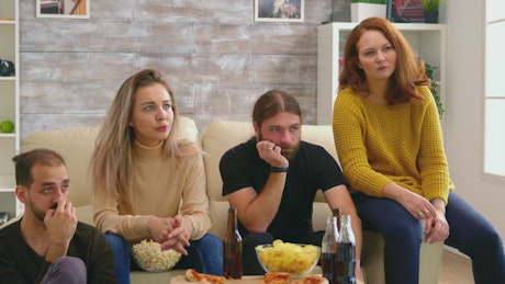 Friends show fear while watching movie on sofa with pizza