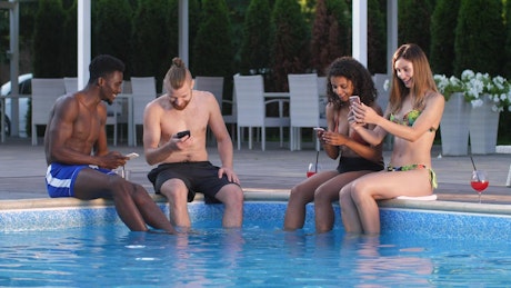 Friends with cell phones sitting on a swimming pool.