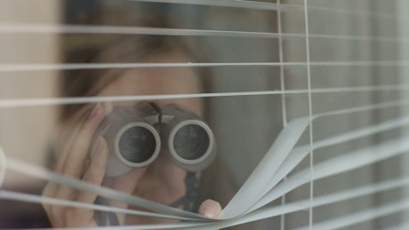 Girl looking through blinds with binoculars from her window.